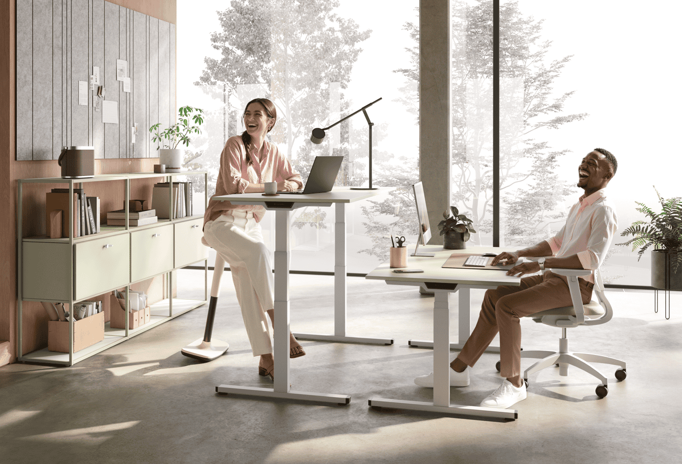 Zwei Personen  in einem modernen, hellen Büro mit ergonomischen Steh-Sitz-Tischen und einer stylischen Regalwand.